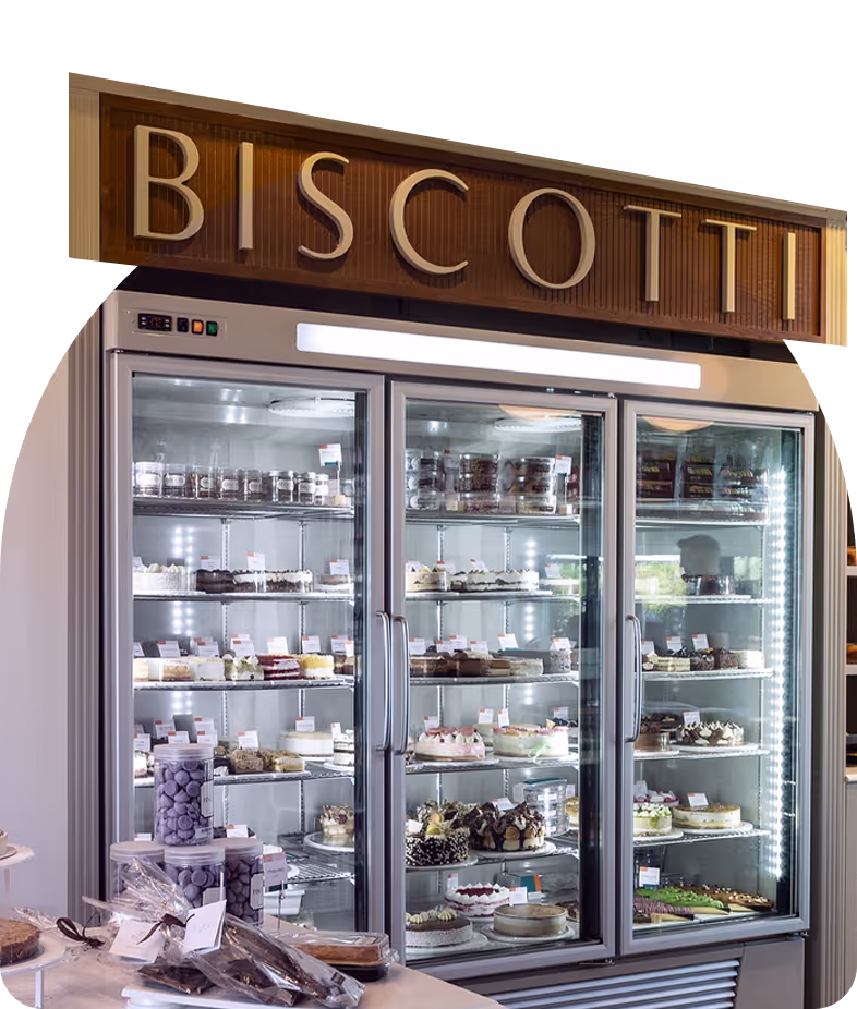Refrigerated display case filled with various cakes, desserts, and packaged biscotti under a wooden sign reading 'BISCOTTI'.