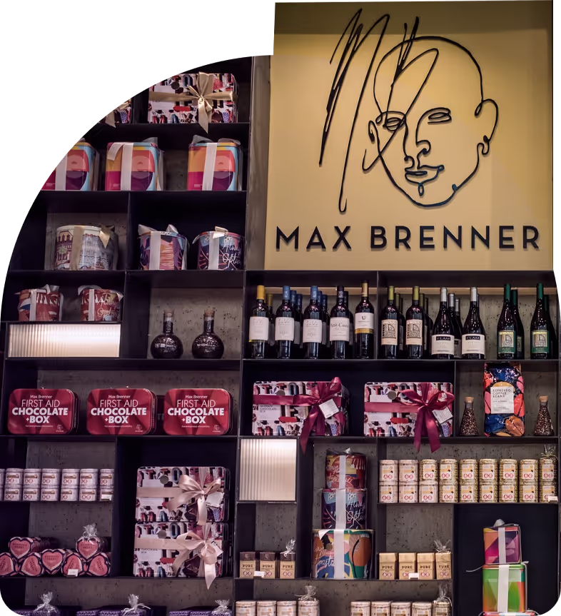 Store display wall with Max Brenner logo above shelves filled with chocolates, gift boxes, wine bottles, and packaged treats.
