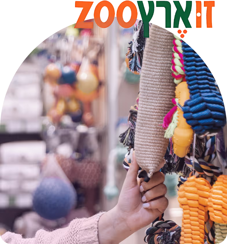 Person's hand holding a beige woven dog chew toy among various colorful dog toys hanging in a pet store.
