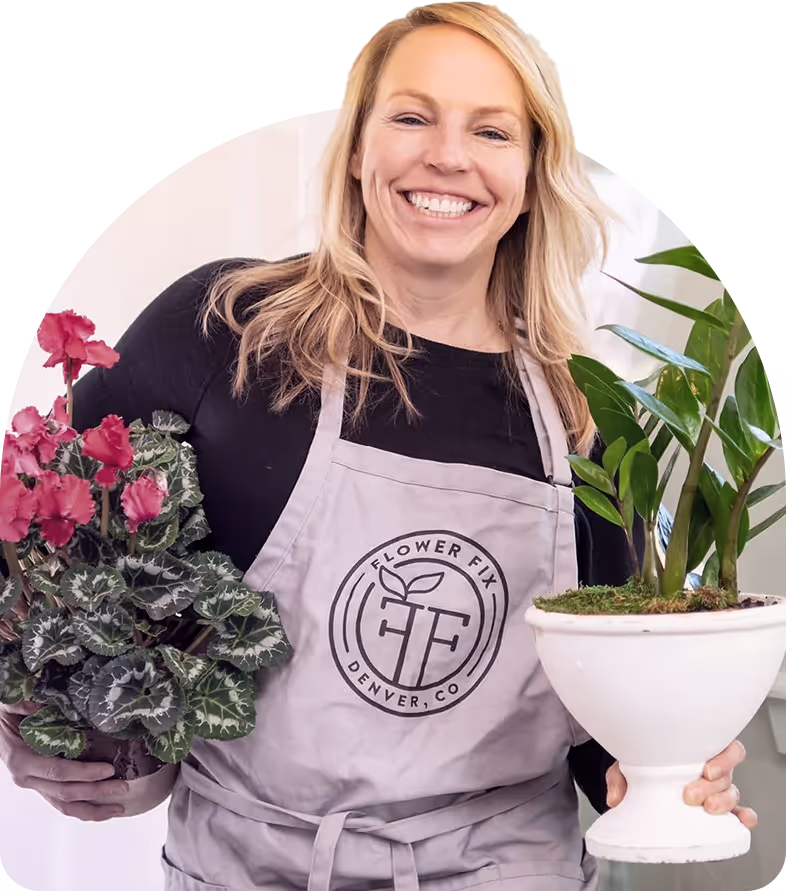 Smiling woman wearing a Flower Fix Denver apron holding a pink flowering plant and a green leafy plant in white pots.